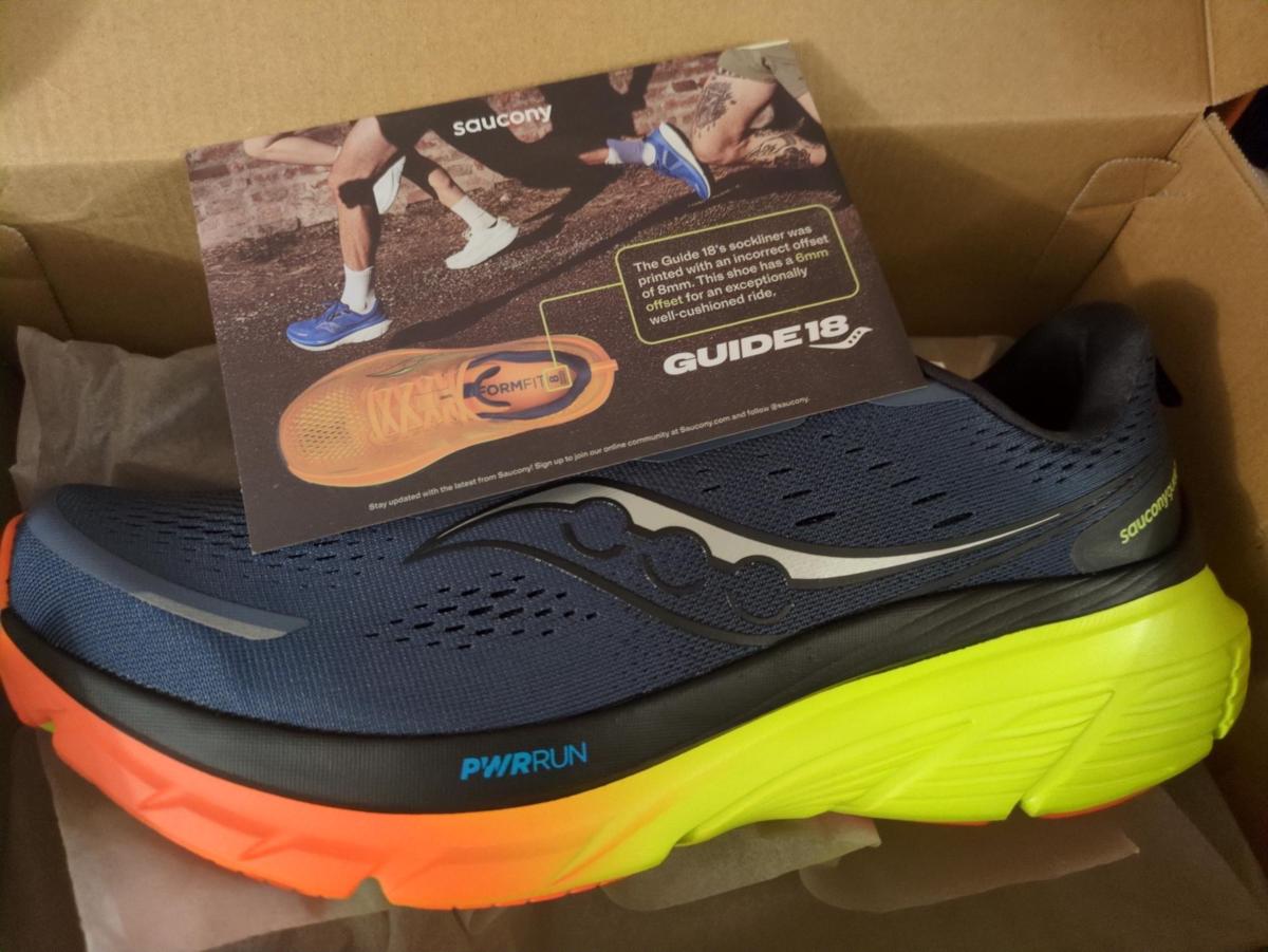A pair of trainers fresh out of the box, along with a piece of card that reads 'The Guide 18's sockliner was printed with an incorrect offset of 8mm. This shoe has a 6mm offset for an exceptionally well-cushioned ride.