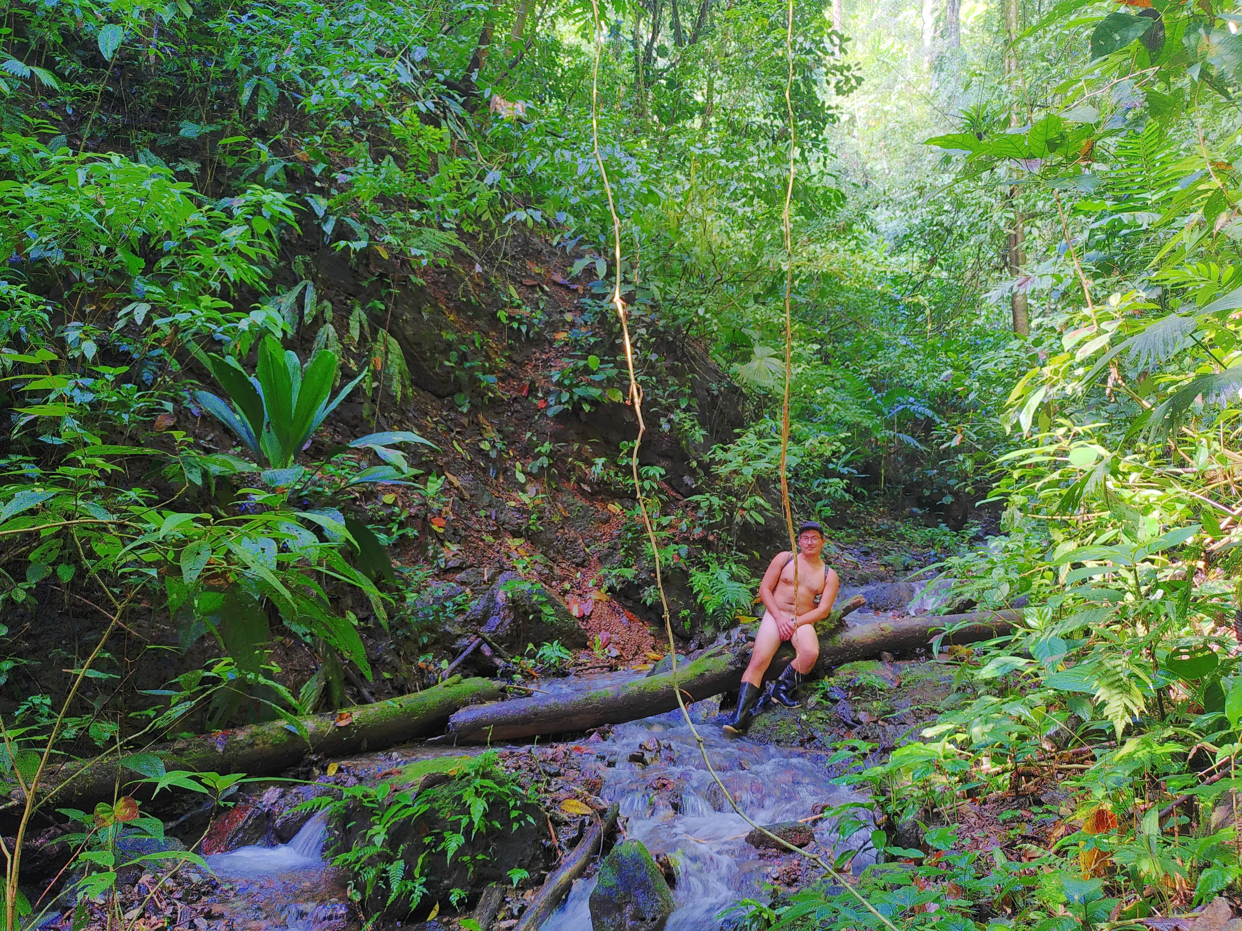 A naked man sitting on a log over a jungle river