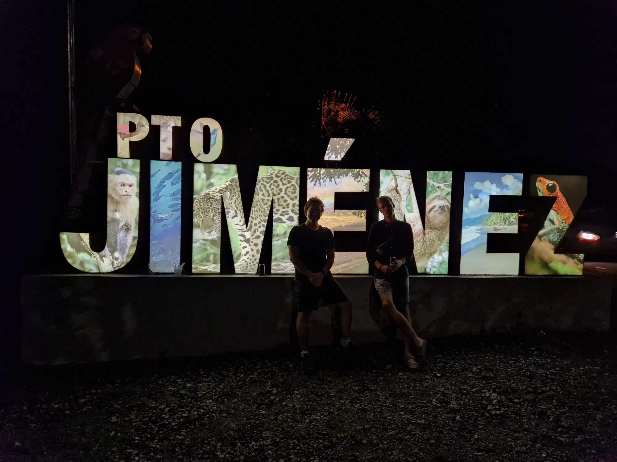 Two men standing in front of an illuminated sign reading ‘Pto Jiménez’, at night