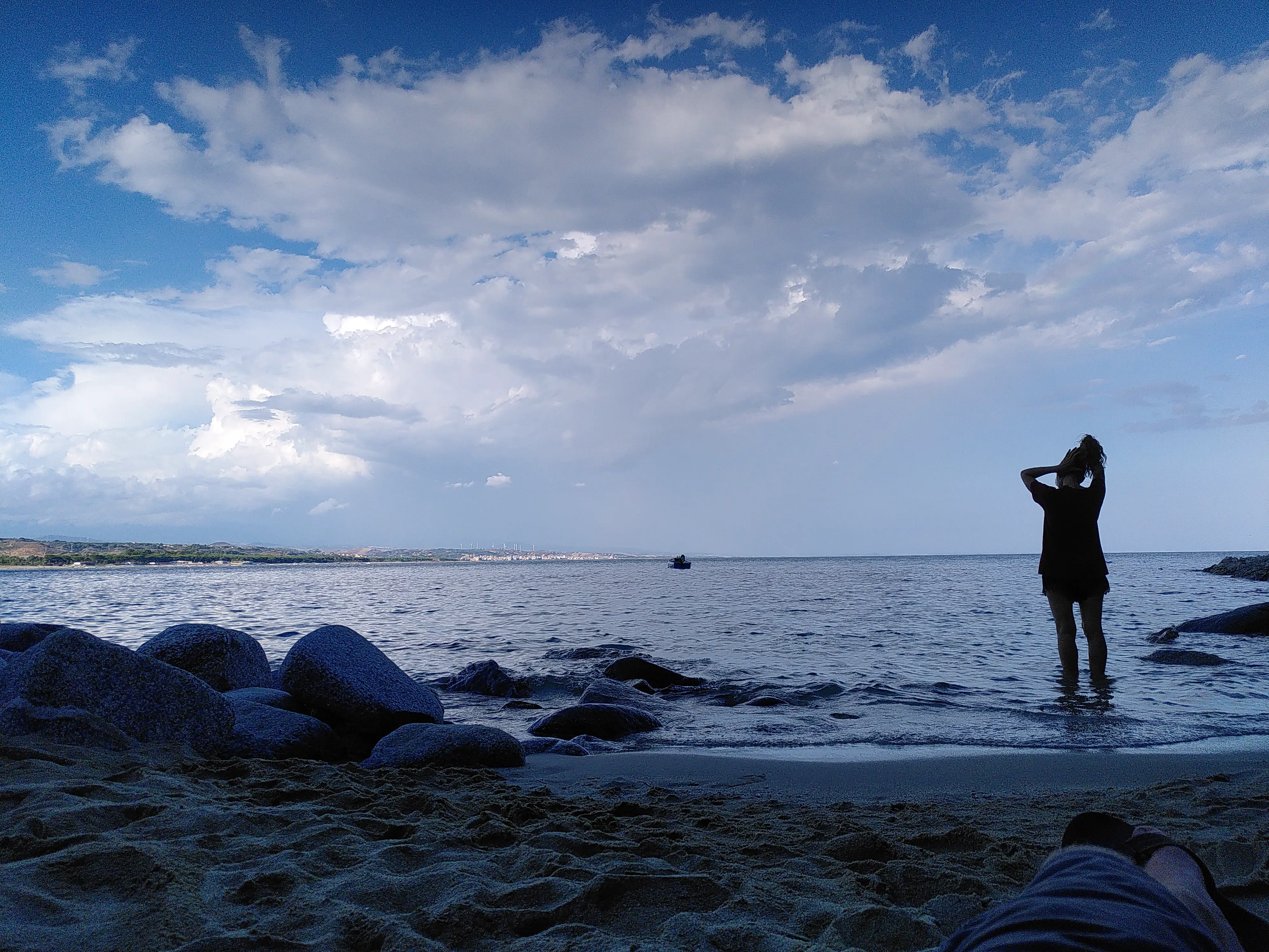 A woman wading in water up to her calves and fixing her hair, seen from behind and in silhouette