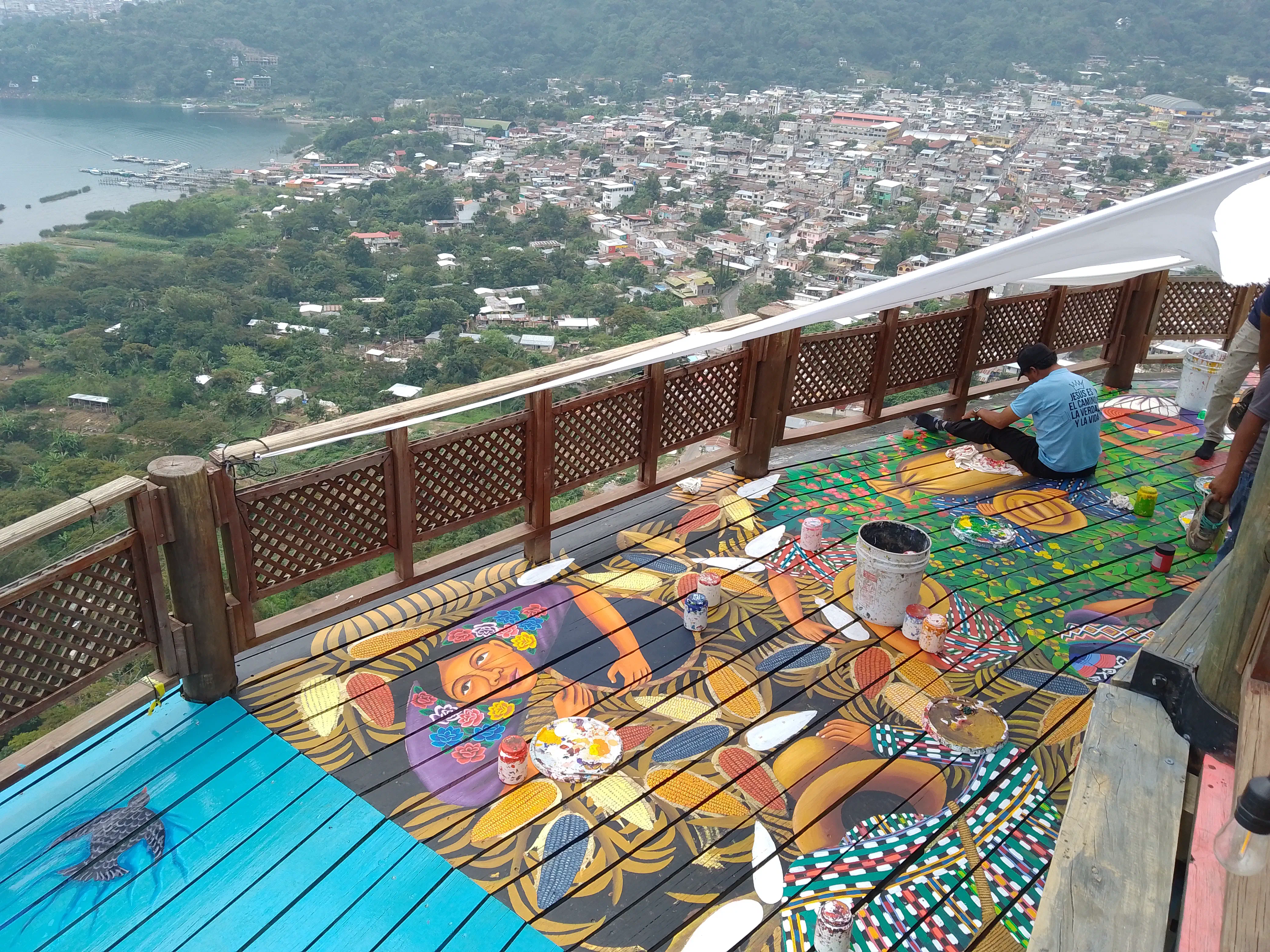 Men painting a mural onto a wooden walkway overhanging a city