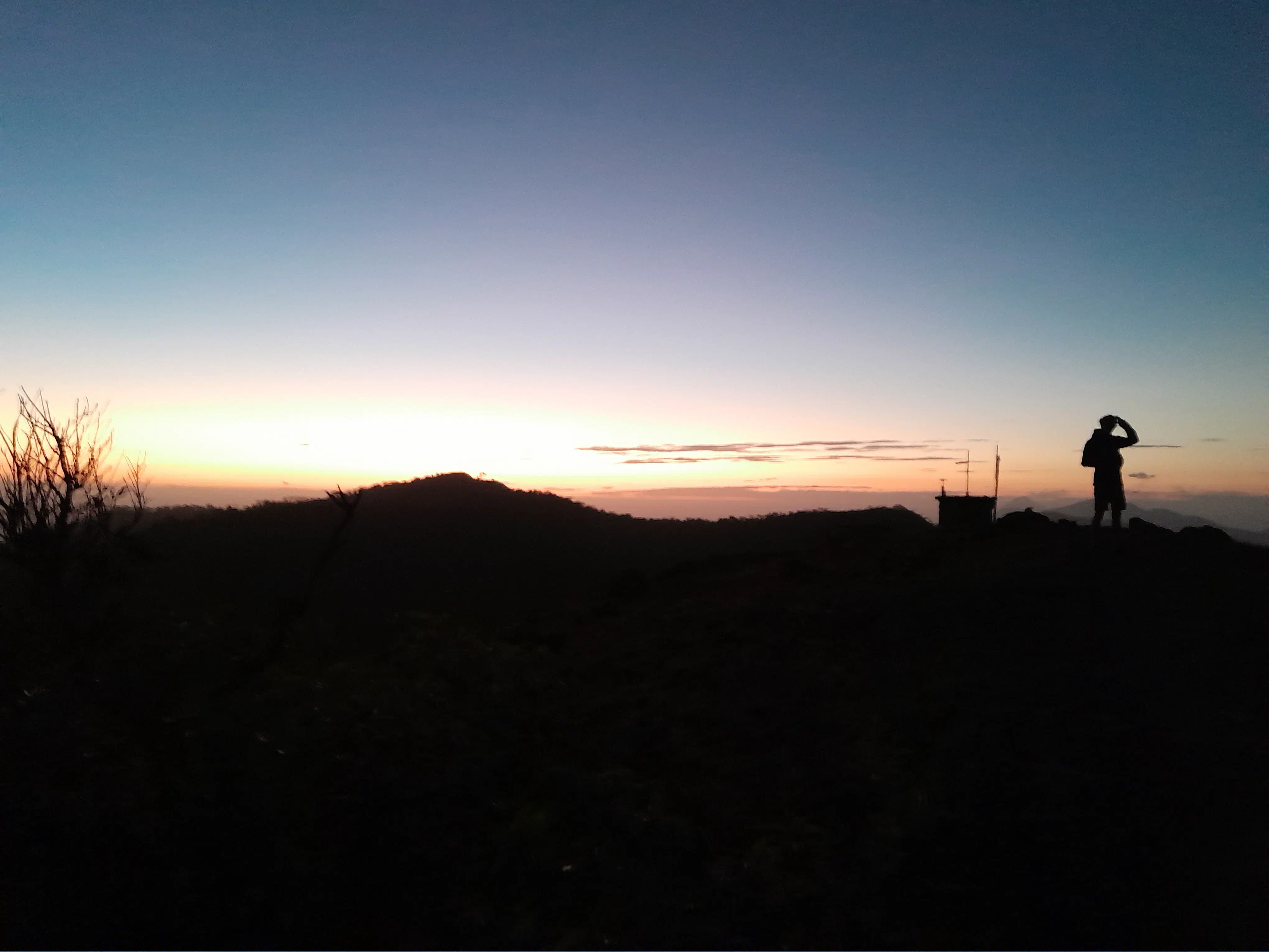 A man stands beside a small structure covered in measuring devices, both silhouetted as the sun sets