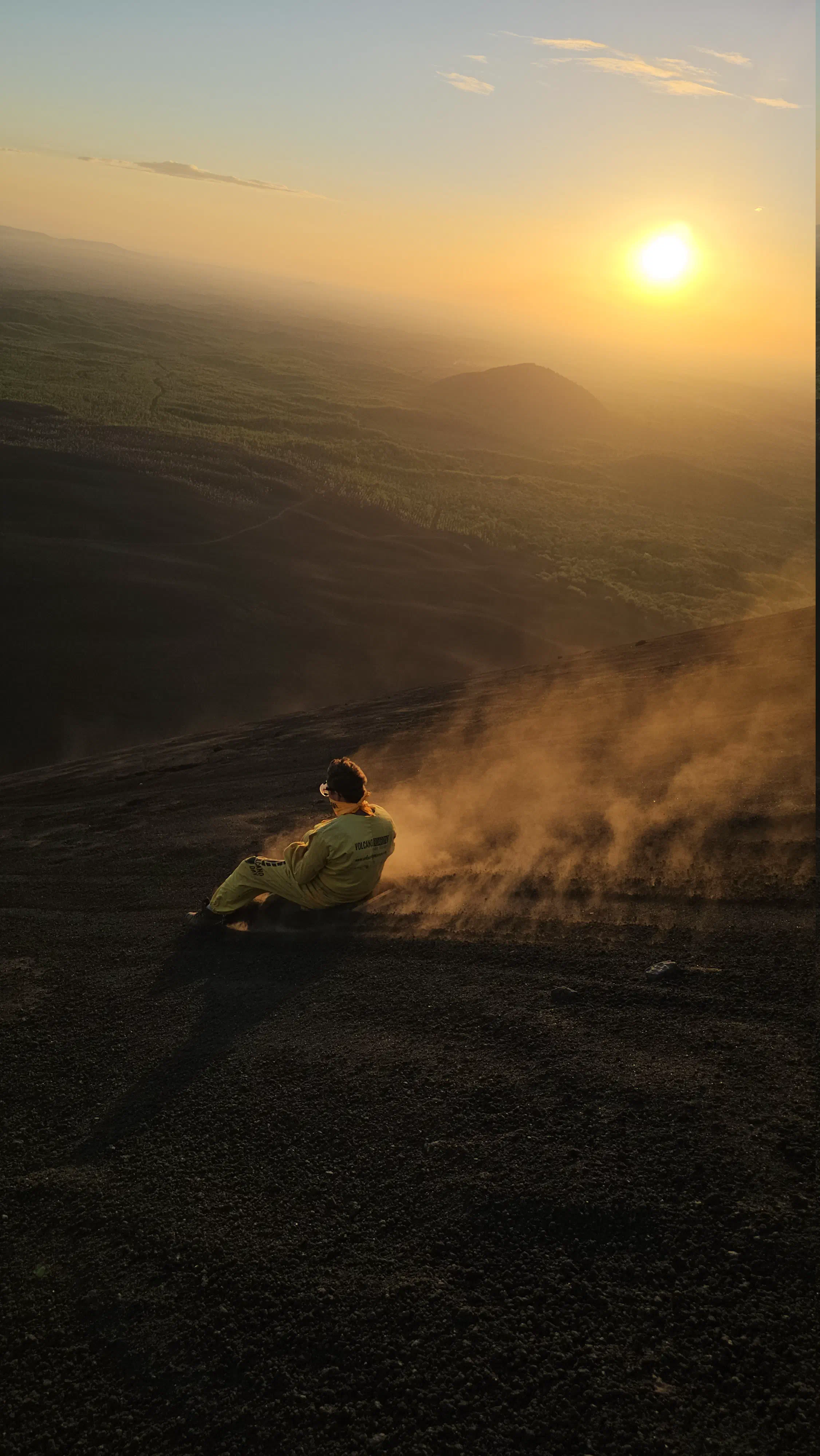 The author sliding down the side of a volcano on a wooden board as the sun sets in the background