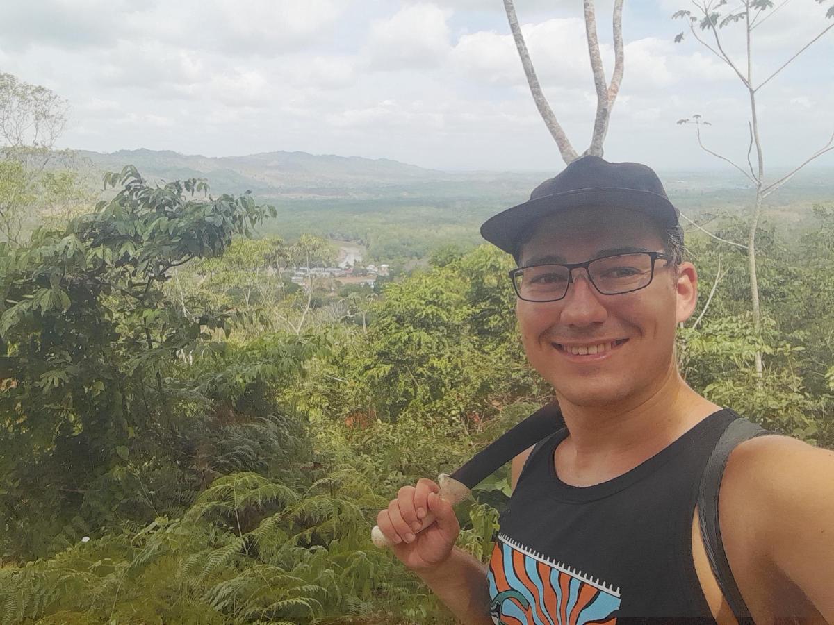 The author in the foreground, resting a machete on his shoulder, with a small village surrounded by jungle in the background