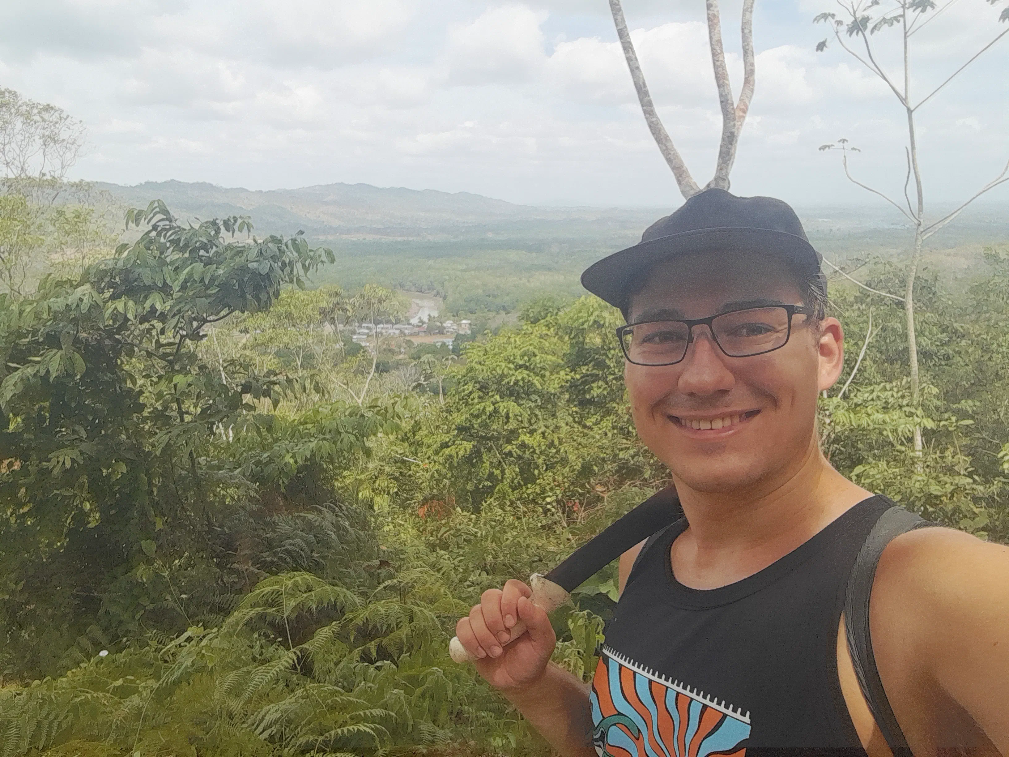 The author in the foreground, resting a machete on his shoulder, with a small village surrounded by jungle in the background