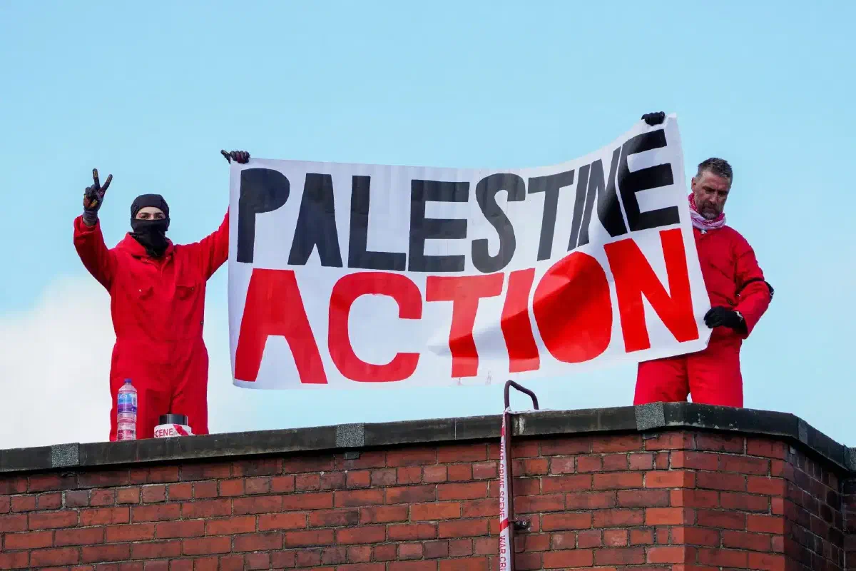 Palestine Action members in black masks and red jumpsuits protest with a banner on top of the roof of a factory