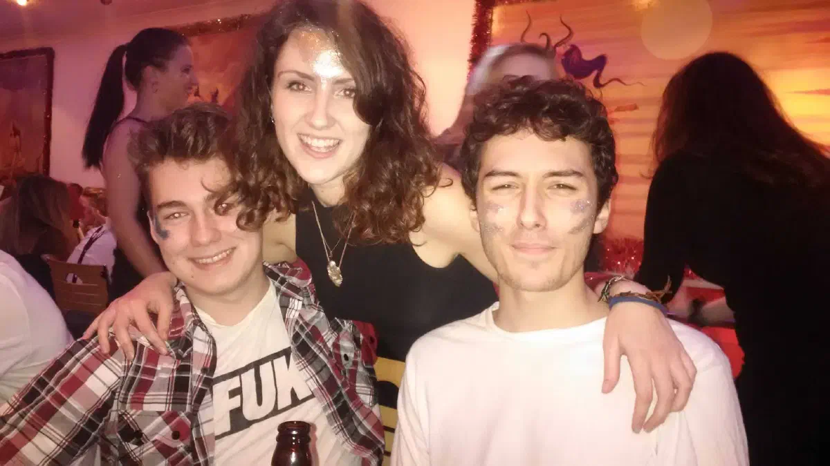 Two men sat at a table with a woman standing behind them, all smiling