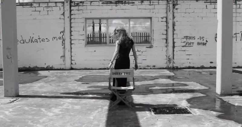 A black and white photo of a woman standing in front of a chair labelled 'Director', in a derelict building with graffiti reading 'who edukates[sic] me?' and 'this is art baby'