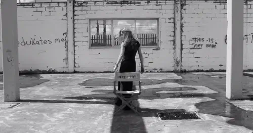 A black and white photo of a woman standing in front of a chair labelled 'Director', in a derelict building with graffiti reading 'who edukates[sic] me?' and 'this is art baby'